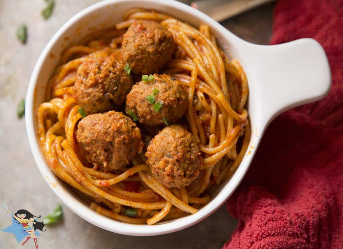 A white bowl of Spaghetti and Meatballs on a table with a red cloth next to it.