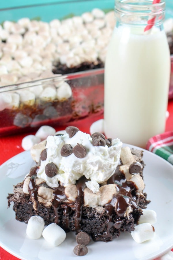 a slice of Hot Cocoa Chocolate Dump Cake on a white plate topped with whipped cream and chocolate chips with a glass of milk and the rest of the cake in the background