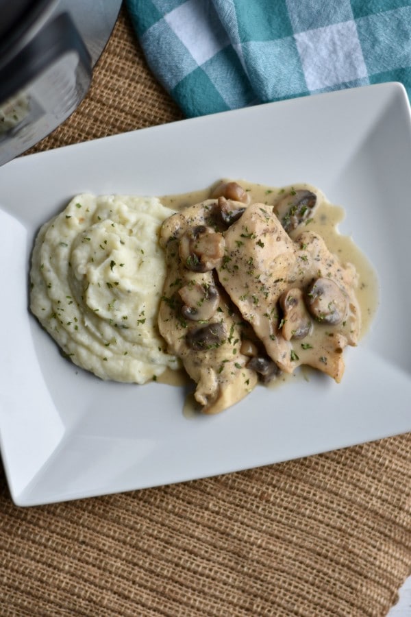 chicken topped with mushrooms and sauce next to mashed potatoes on a white plate with an instant pot and blue cloth in the background