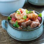 A ceramic bowl brimming with an Instant Pot Shrimp and Sausage Gumbo creation features rice crowned with succulent shrimp, savory meat, diced vegetables, and a garnish of green onions on a wooden surface.