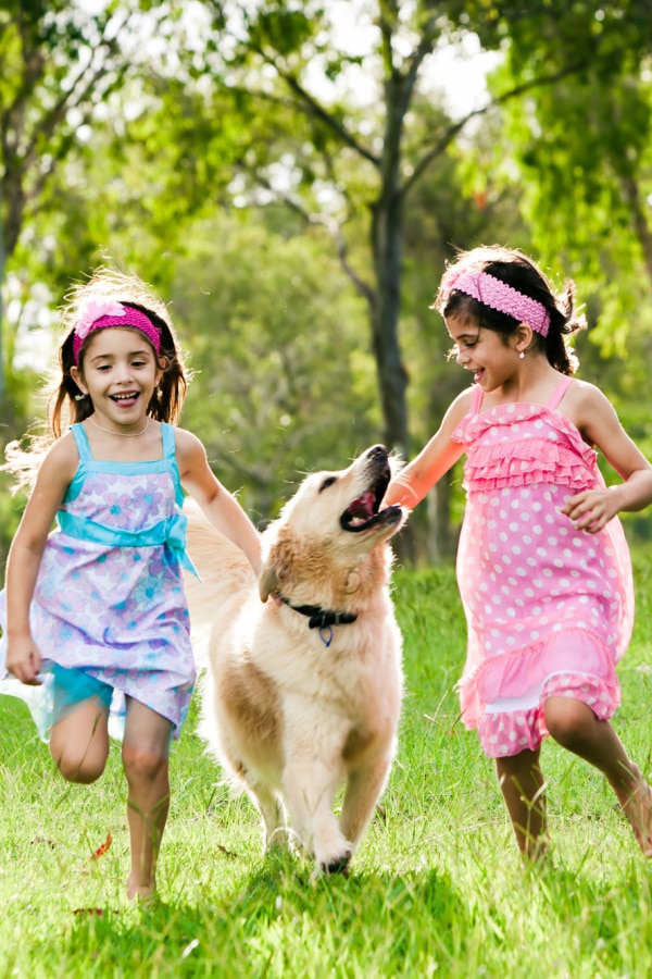 two girls running with their dog outside on grass with trees in the background