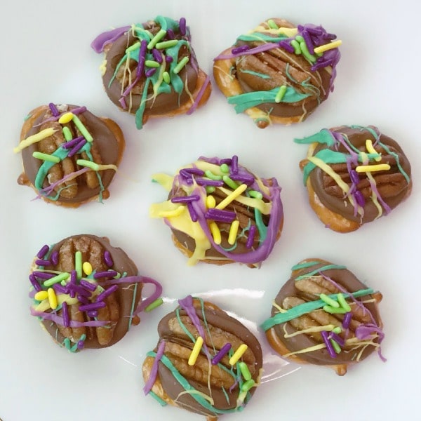 pretzels topped with chocolate, a pecan, purple frosting and colored sprinkles, all on a white counter