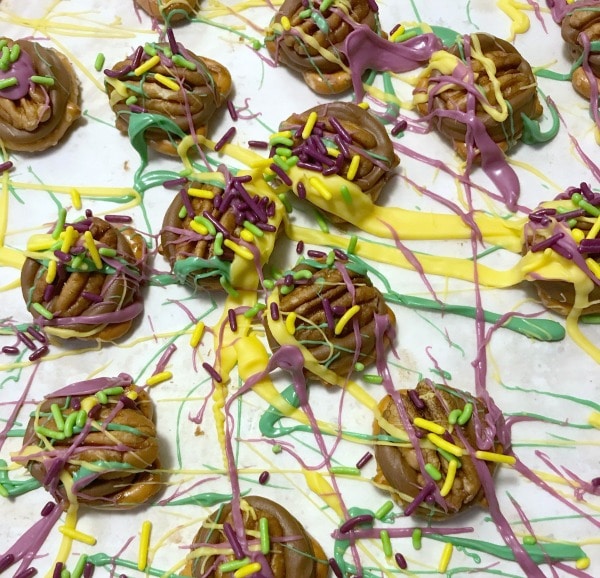 pretzels topped with chocolate, a pecan, purple and yellow frosting and colored sprinkles, all on a white paper