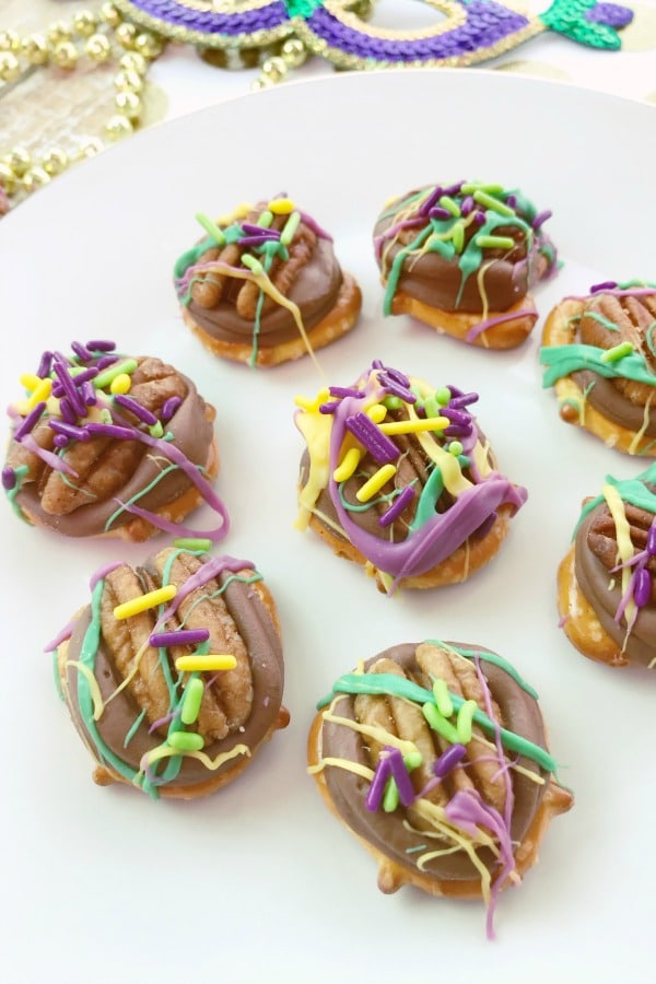 pretzels topped with chocolate, a pecan, purple frosting and colored sprinkles, all on a white plate
