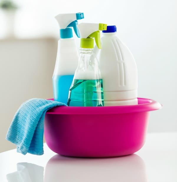a pink bowl with two spray bottles, one plastic jug, and a blue rag in it, all on a white table