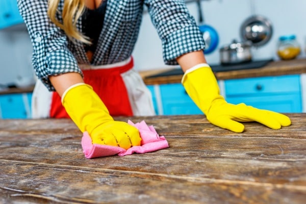 a lady wearing yellow gloves cleaning a wood table with a pink rag with the rest of the kitchen in the background