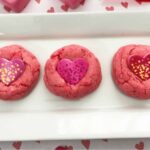 Three pink cookies made from strawberry cake mix with heart-shaped decorations are arranged in a row on a white rectangular plate.
