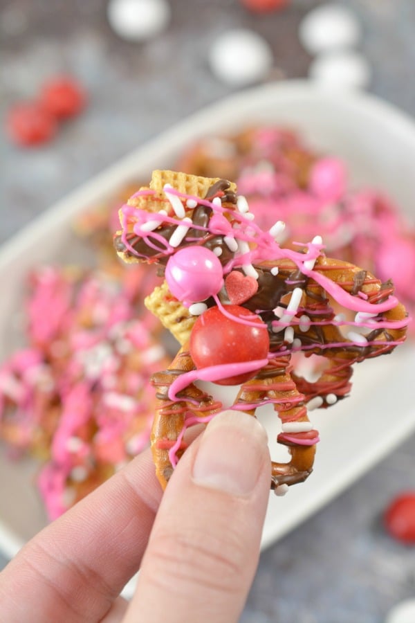 a hand holding a piece of Valentine Bark, which is a mix of chex, pretzels, melted chocolate, white sprinkles, and pink and white candy