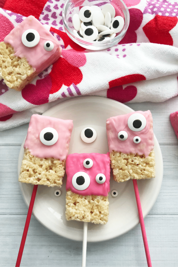 Valentine Rice Krispie Bars with pink frosting and monster candy eyes on a plate with more in the background on a heart cloth