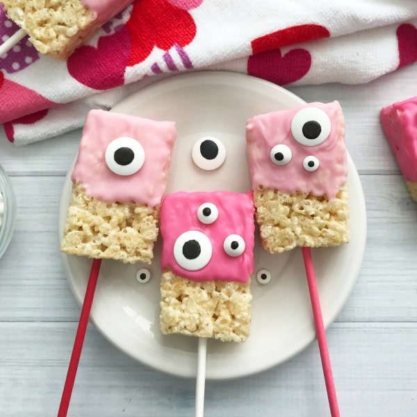 Valentine Rice Krispie Bars with pink frosting and monster candy eyes on a plate with more in the background on a heart cloth