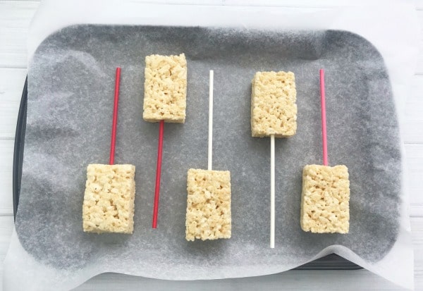 a tray lined with parchment paper with rice crispie treats with sticks in them.