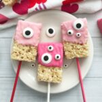 Three Valentine's Day Rice Krispie Treats on sticks, half-dipped in pink icing with googly eye decorations, placed on a white plate.
