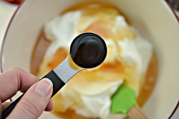 a hand holding a measuring spoon of vanilla over a bowl of yogurt and honey