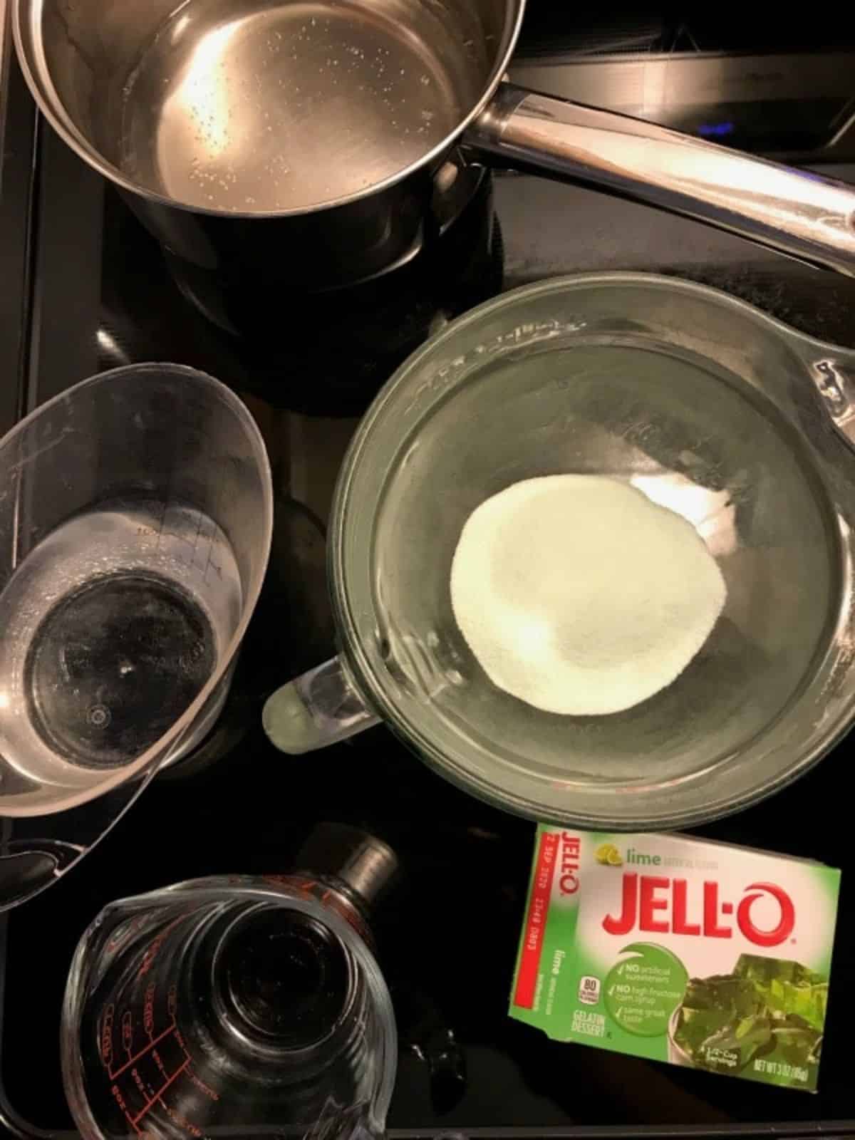 measuring cups and pans full of water and jello, and a box of green jello.