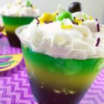 A delightful layered dessert cup with whipped cream, colorful sprinkles, and candies on top sits perfectly on a purple zigzag-patterned surface, showcasing its vibrant and inviting appearance.