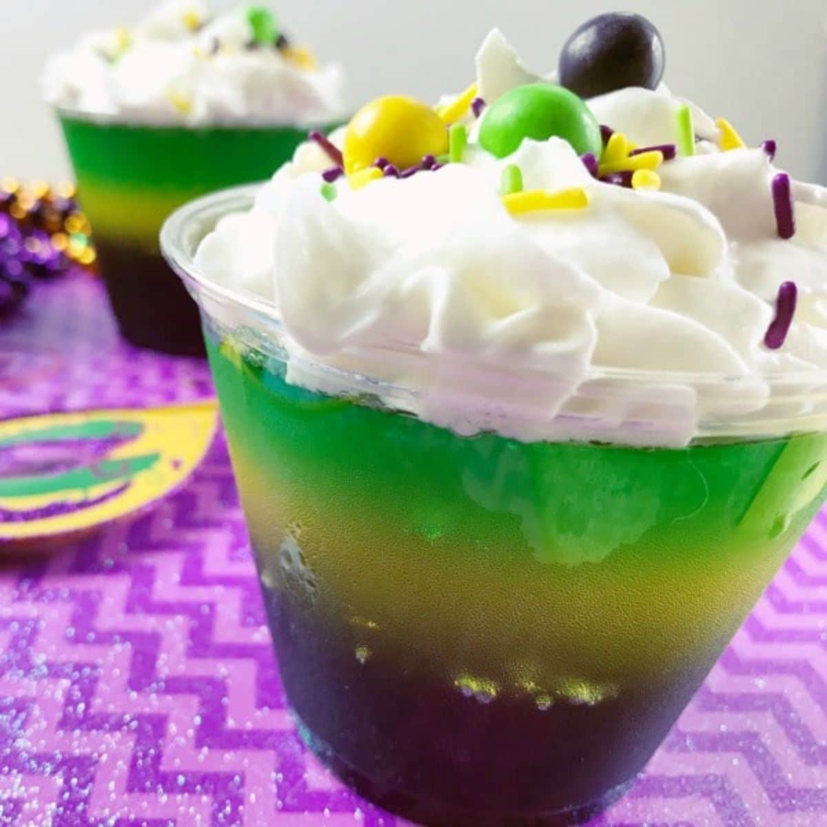 Layered Jello for Mardi Gras in cups topped with whipped cream, sprinkles and skittles on purple paper.