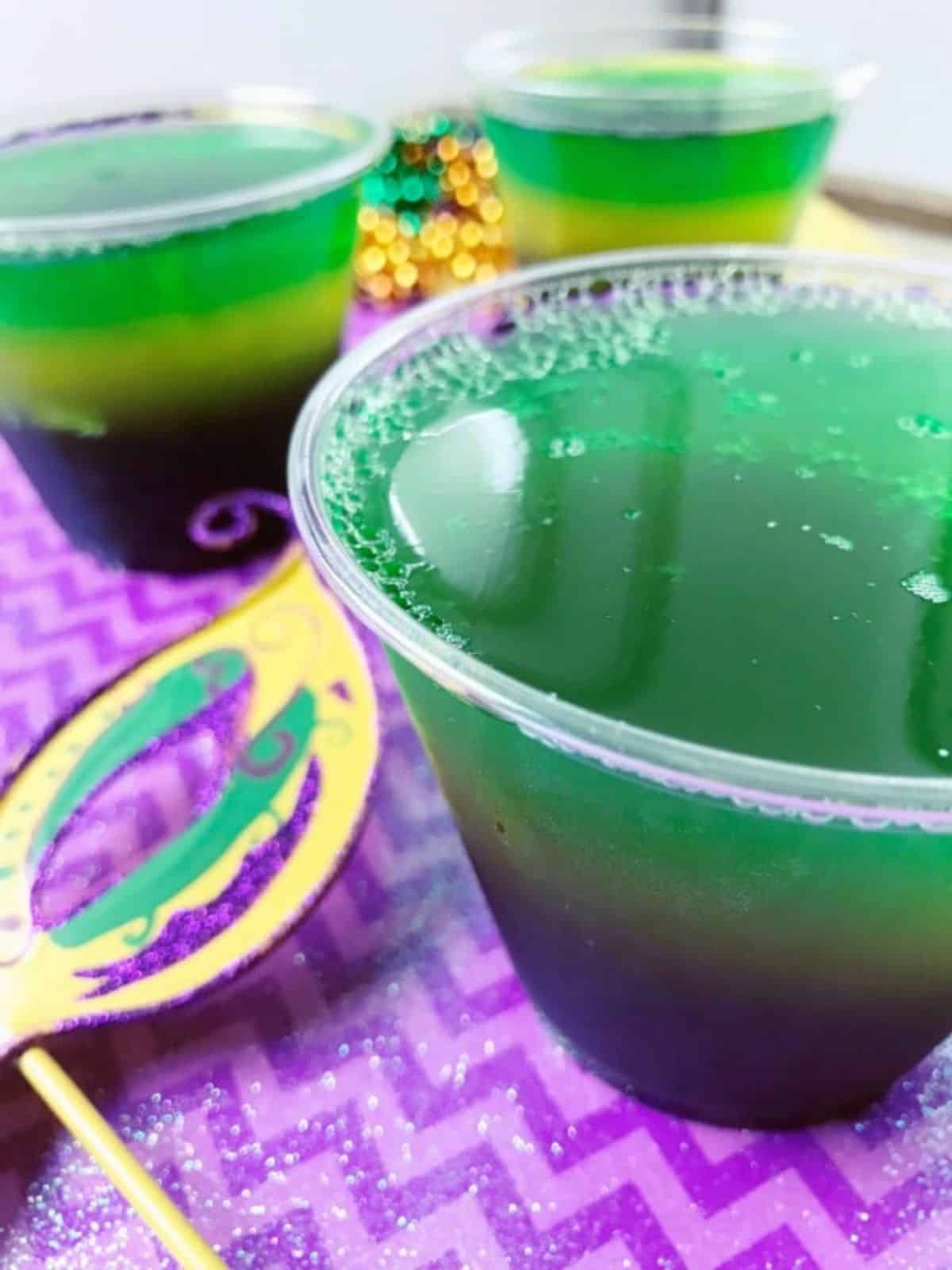Layered Jello for Mardi Gras in cups on purple paper. Mardi Gras mask on the side.