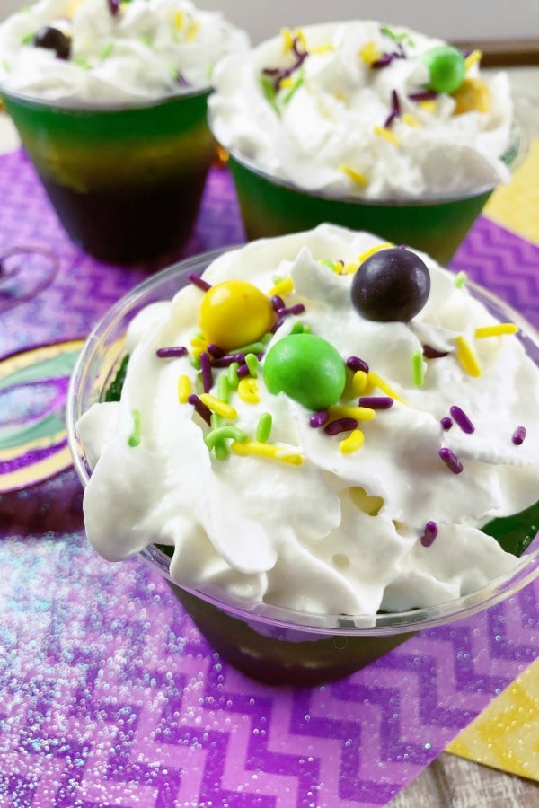 Mardi Gras Layered Jello in cups topped with whipped cream, sprinkles and skittles on purple and yellow paper