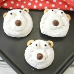 Three bear-themed cookies, reminiscent of polar bear donuts, with white frosting, chocolate chips for noses, and pretzel pieces for ears are arranged on a black surface.