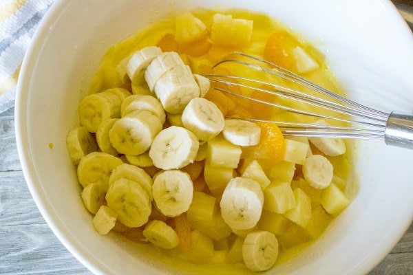 bananas, pineapple chunks, mandarin oranges, and pudding in a white mixing bowl with a wire whisk in it