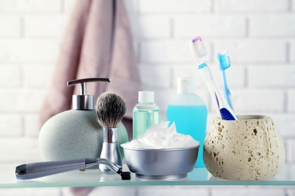 a glass counter with shaving cream, razor, soap dispenser, mouthwash, and toothbrush holder with toothbrushes with a towel on a brick wall in the background
