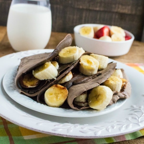 Chocolate Nutella and Banana Crepes on a white plate on a table with a glass of milk and a bowl of bananas and strawberries in the background