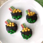 Four Oreo St. Patrick's Day treats resembling pots of gold on a white plate. Each has gold candy, green clover decorations, and a rainbow candy strip.