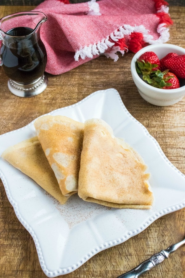 Snickerdoodle Crepes on a white plate with a glass jar of syrup, a white bowl of strawberries, and a red cloth in the background