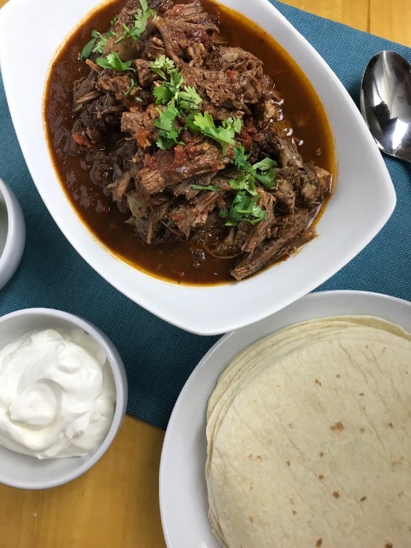 carne guisada in a white dish next to a spoon, a white bowl of sour cream and a white plate with tortillas on it