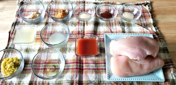 raw chicken breasts, glass bowls with the seasonings and other ingredients needed for chicken enchilada tacos all on a cloth on a table