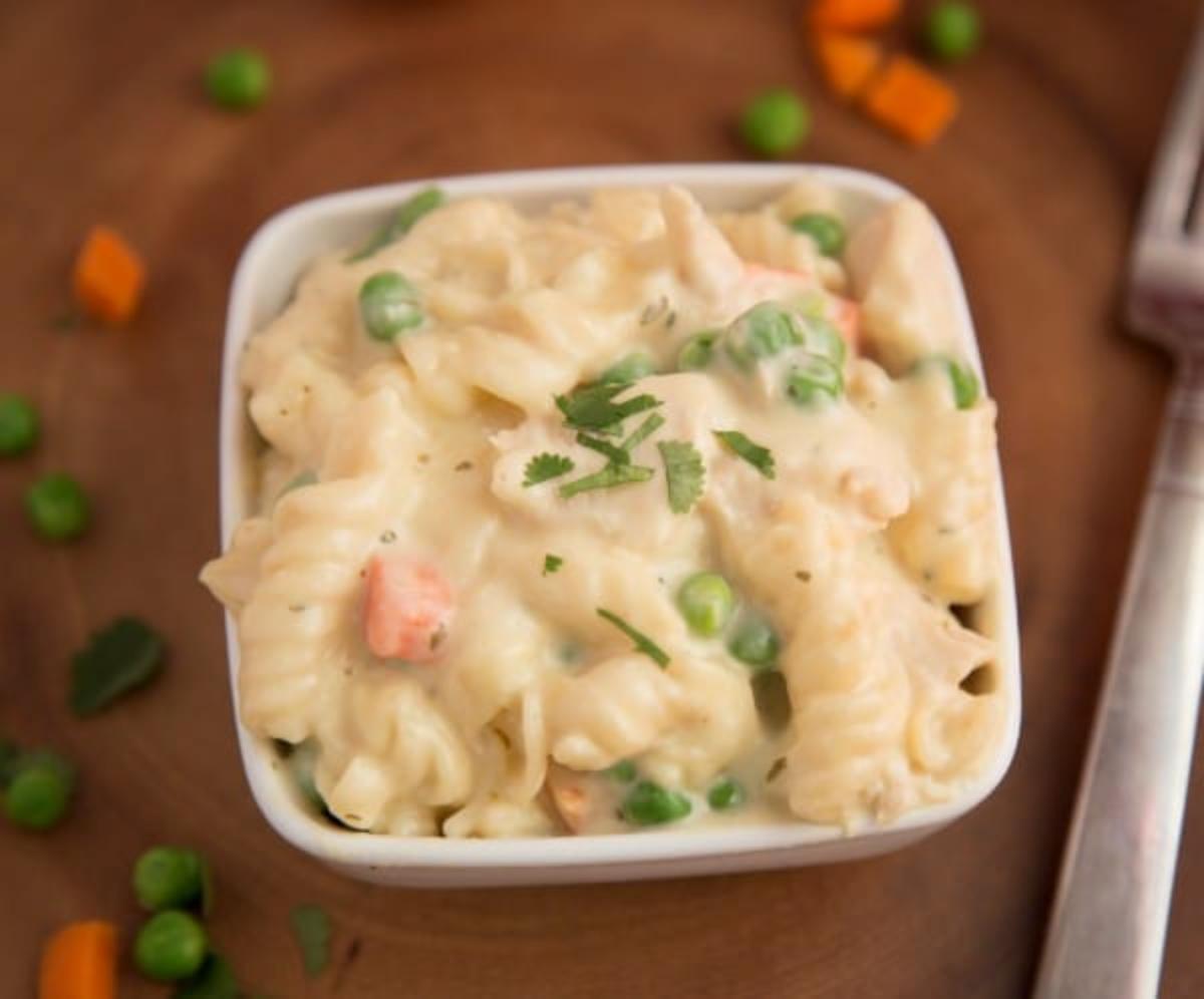 Creamy chicken pasta in a white bowl on a tale with peas & carrots and a fork in the background.