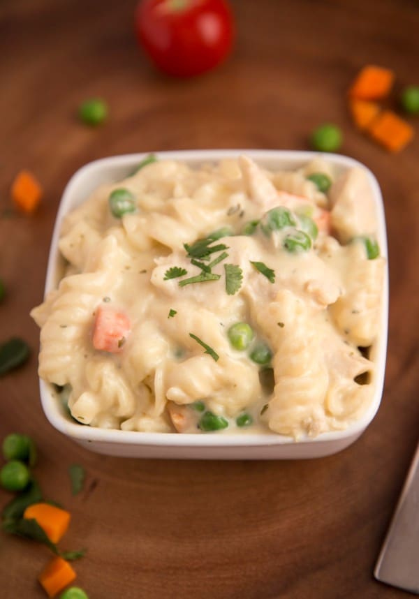 creamy chicken pasta in a white bowl on a table with peas and carrots and an apple in the background