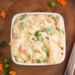 A square white bowl filled with instant pot creamy chicken pasta, mixed with peas and diced carrots, garnished with chopped herbs, placed on a wooden surface.