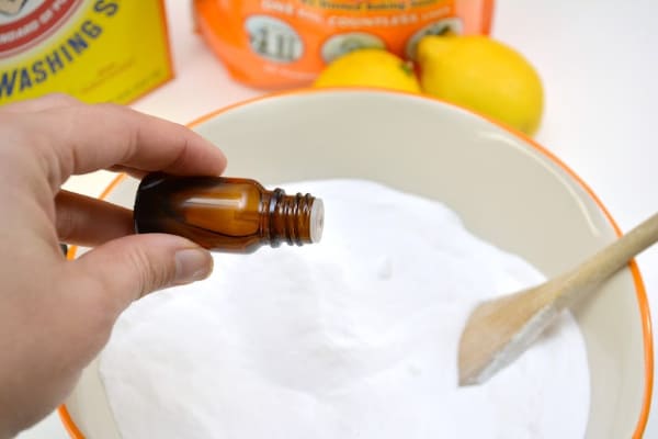 a hand pouring essential oil into a white bowl of washing soda and baking soda in a white bowl with a wooden spoon 