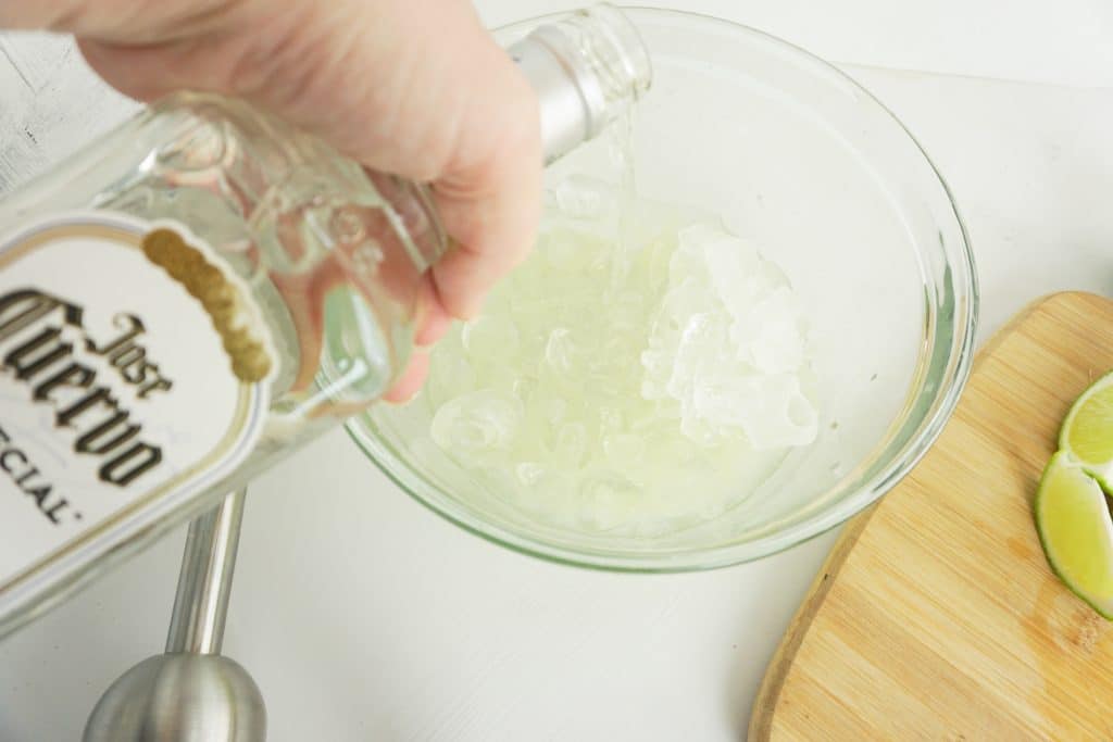 a hand pouring tequila over a glass bowl of ice