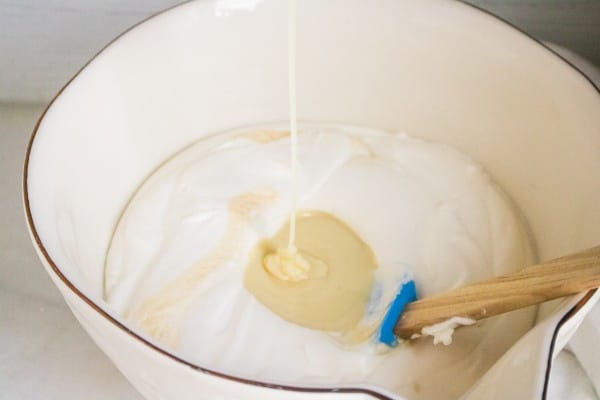 sweetened condensed milk being poured into a white bowl of heavy cream with a spatula in it