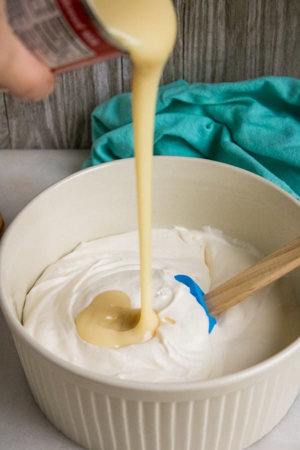 condensed milk being poured into an off white bowl of whipped heavy cream with a spatula in it