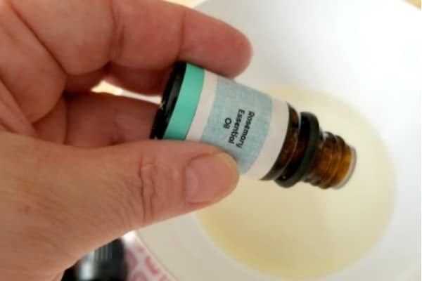 a lady's hand holding a bottle of rosemary essential oil over a bowl of liquid