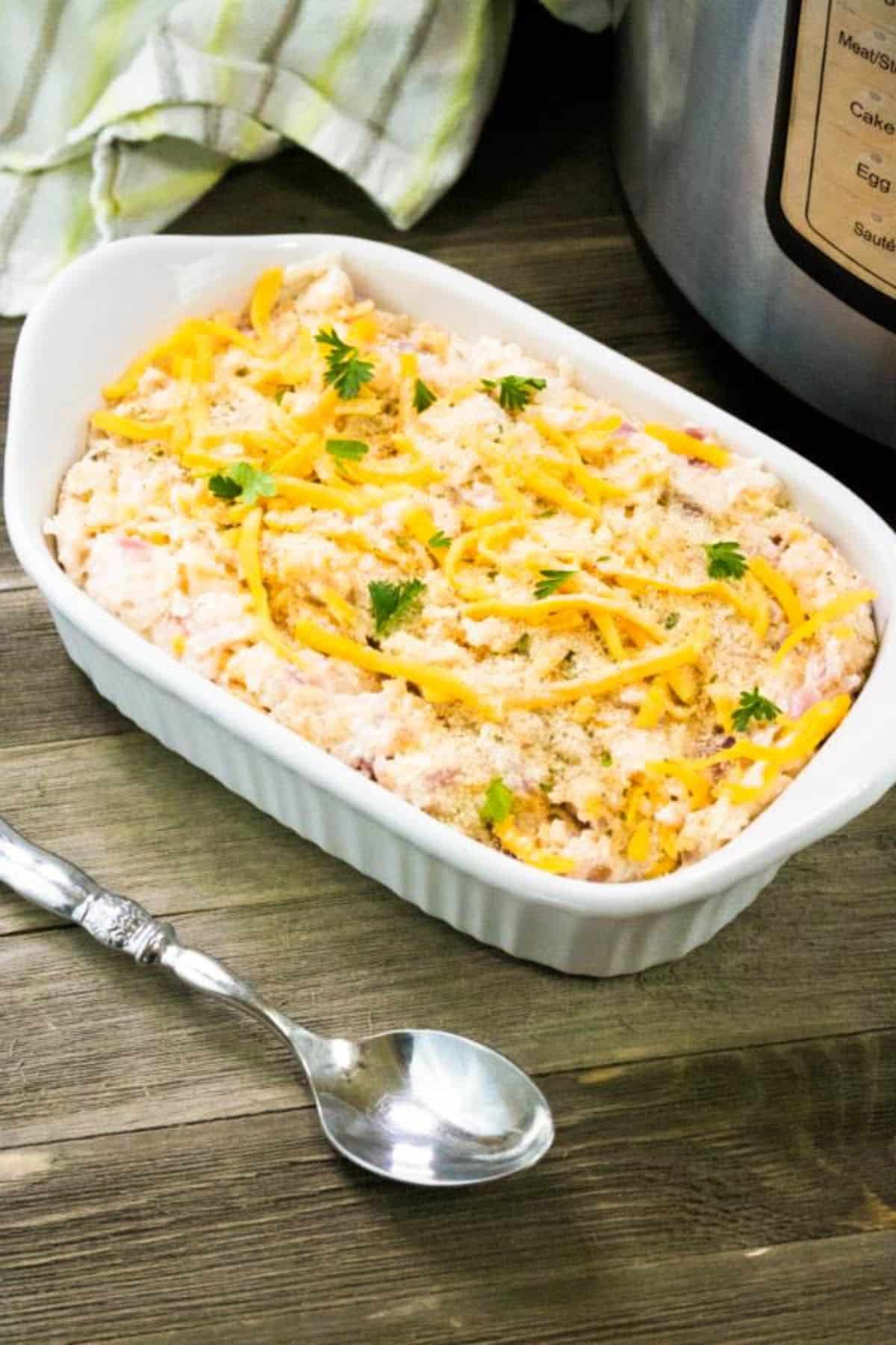 Instant Pot Cheesy Potatoes in a white baking dish on table next to Instant Pot and serving spoon.