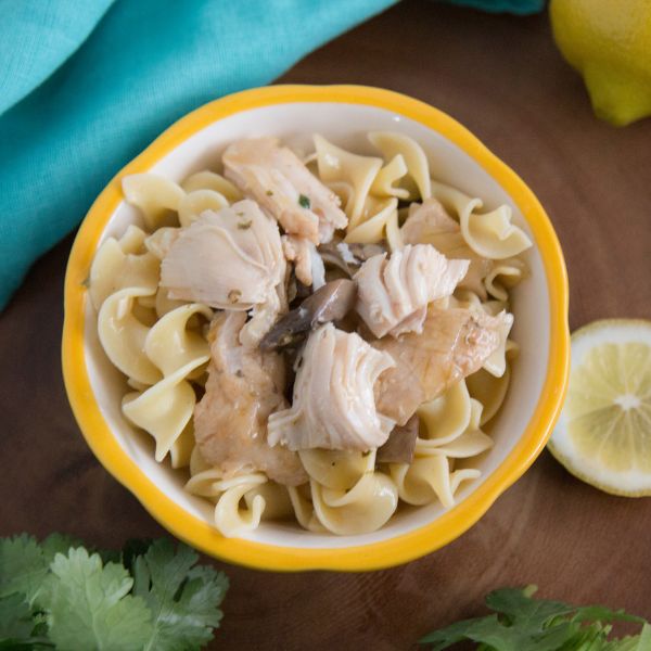 a bowl of noddles topped with lemon chicken and mushrooms with a blue cloth and lemon in the background