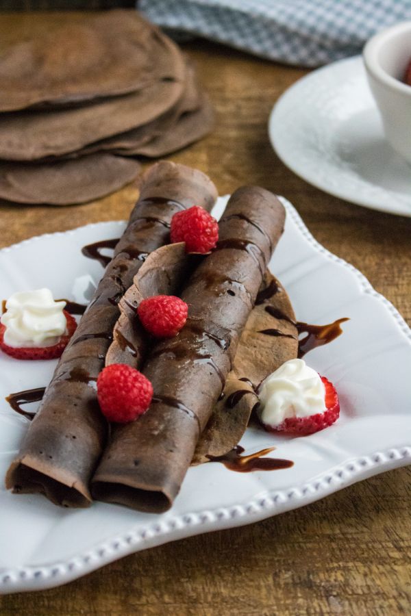 Chocolate Crepes on a white plate topped with raspberries with more crepes on the table in the background