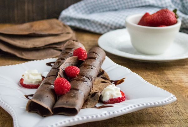 Chocolate Crepes on a white plate topped with raspberries with more crepes on the table and a white cup on a plate filled with strawberries in the background