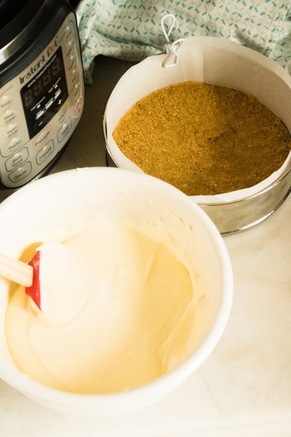 a mixing bowl full of the orange cream batter for the cheesecake next to a springform pan filled with a crust next to an instant pot