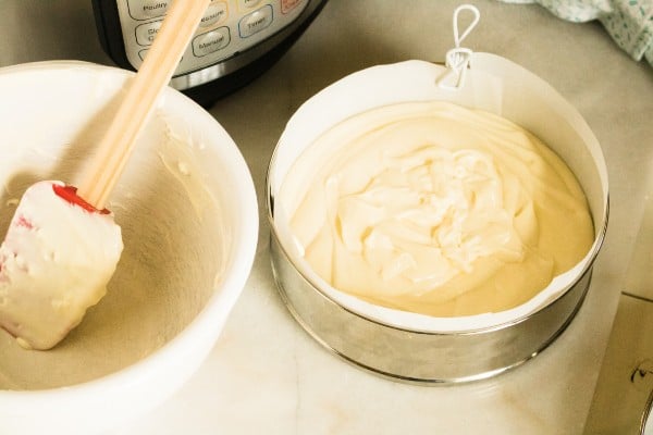 an empty mixing bowl with a spatula in it next to a springform pan filled with orange cream cheesecake batter with an instant pot in the background