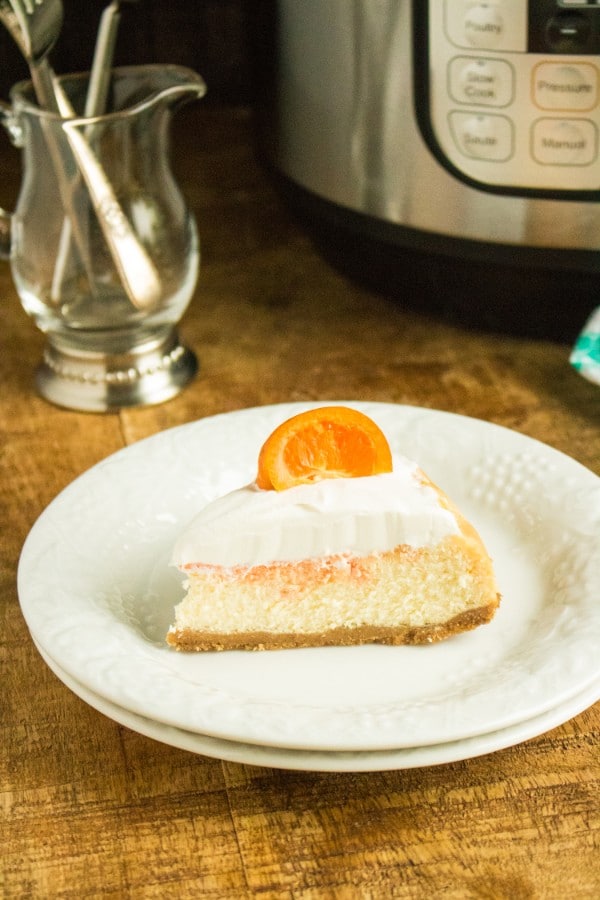 Slice of orange cream cheesecake with a mandarin slice garnish on plate on table with glass of utensils and Instant pot in background