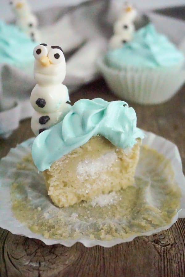 a cupcake cut open showing sprinkles hidden inside with Olaf character cupcake toppers on a wood table with a grey and white linen in the background