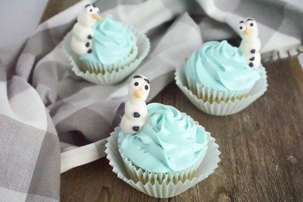 three cupcakes decorated with blue icing and a snowman resembling DIsney's Frozen Olaf character on top of the cupcake on a wood table with a grey and white linen in the background