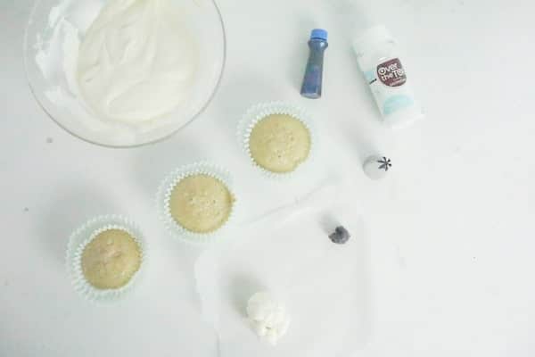 a bowl of white frosting, three cupcakes, white and black fondant, decorating tip, blue food coloring and sugar sprinkles on a white table