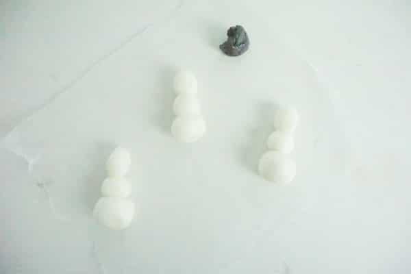 forms of marshmallow fondant snowmen laying on a white table with a blob of black marshmallow fondant