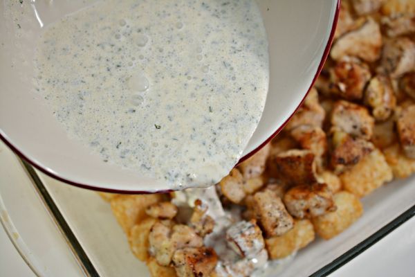 heavy cream and ranch dressing mix being poured from a white bowl into a white casserole dish filled with cooked chicken and tater tots